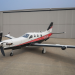 A white and red Daher TBM 910 turboprop parked on the ramp in front of a hangar