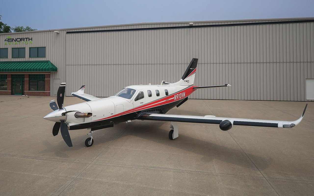A white and red Daher TBM 910 turboprop parked on the ramp in front of a hangar