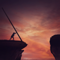 Silhouetted figure at a cliff edge at sunset using a long pole to bridge a gap