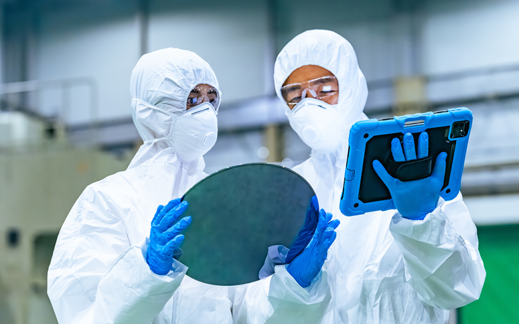 Two workers in cleanroom suits examine a silicon wafer and a tablet