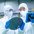 Two workers in cleanroom suits examine a silicon wafer and a tablet